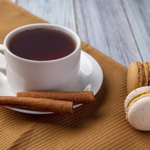 side view cup of tea with cinnamon and macaroons on a brown towel on a gray background